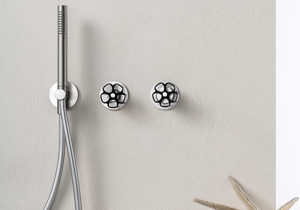 L'immagine rappresenta un set doccia a incasso di design, caratterizzato da un doccino minimale a stelo e due distintive manopole di comando nere a forma di ruota industriale su base in finitura acciaio.